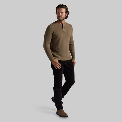 A man modeling a long-sleeve henley sweater in a warm taupe-brown color, paired with black pants and brown boots, against a solid light gray background.