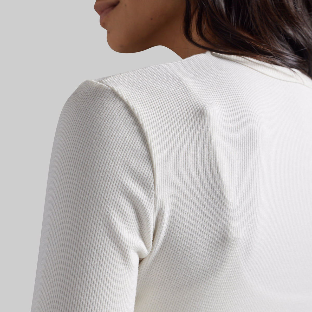 Close-up of a model wearing a ribbed white long-sleeve top, showing the back and shoulder detail against a light gray background.