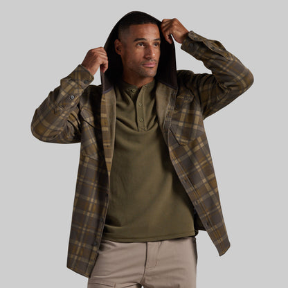 A man wearing a brown and olive green plaid hooded flannel shirt over an olive green henley shirt and khaki pants, posing with the hood up against a solid gray background.