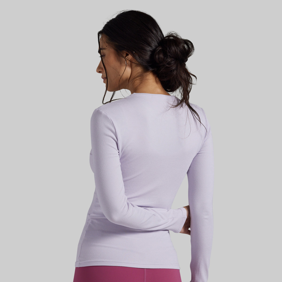 A woman shown from the back wearing a fitted long-sleeve athletic top in a very light lavender-purple color, paired with magenta leggings, against a solid light gray studio background.