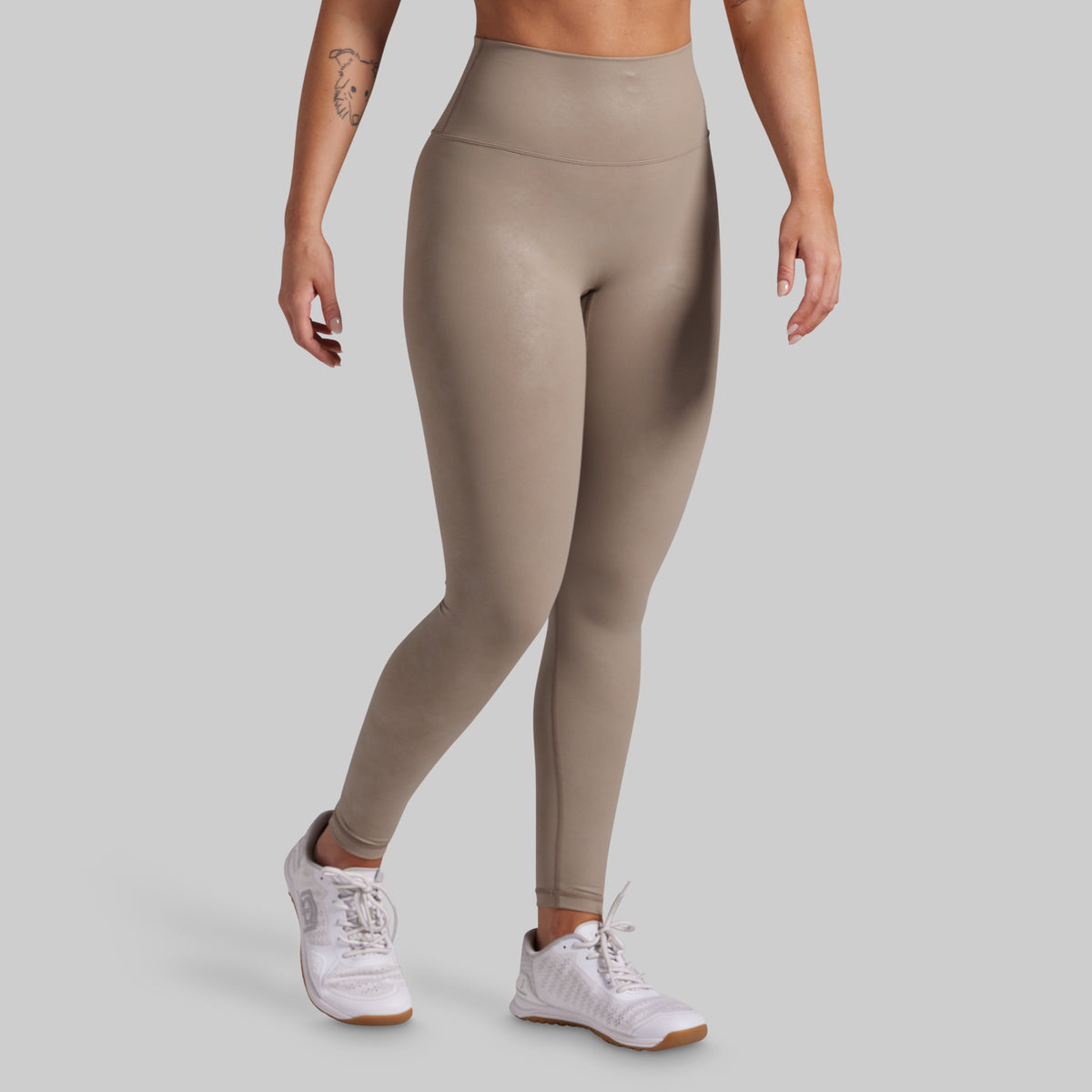 A model wearing high-waisted, full-length athletic leggings in a taupe-beige color, paired with white sneakers, shown from the waist down against a solid light gray background.