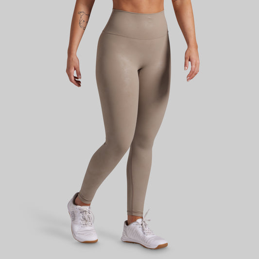 A model wearing high-waisted, full-length athletic leggings in a taupe-beige color, paired with white sneakers, shown from the waist down against a solid light gray background.