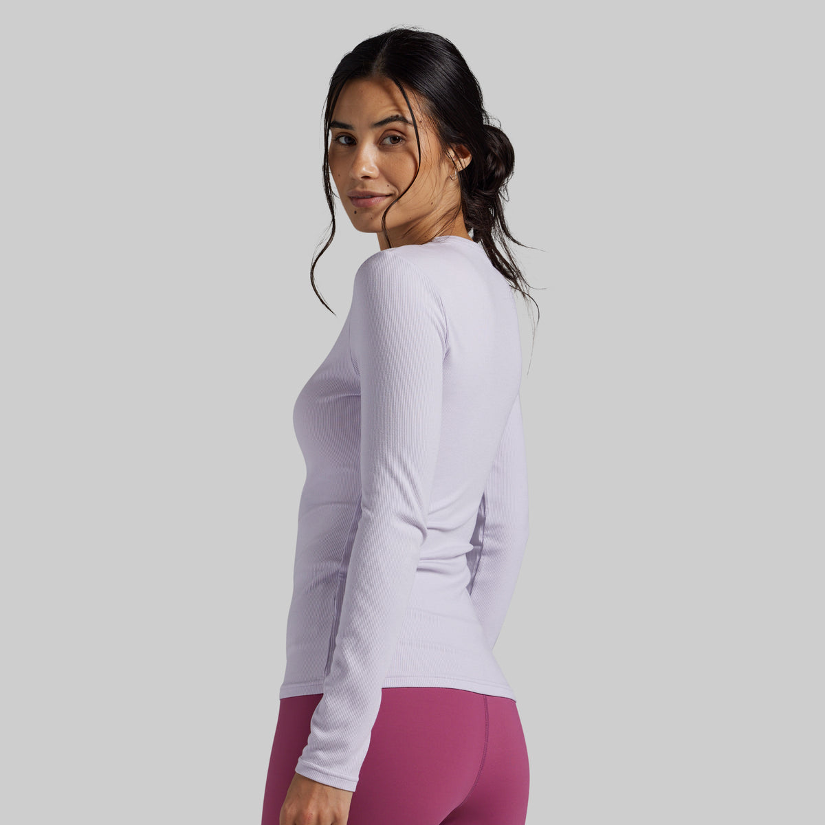 A woman wearing a fitted long-sleeve athletic top in a very light lavender-purple color, paired with magenta leggings, shown from a side-back angle against a solid light gray studio background.