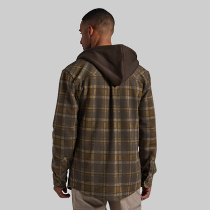 A man viewed from behind wearing a brown and olive green plaid hooded overshirt with a dark brown hood, paired with light khaki pants, against a solid gray background.