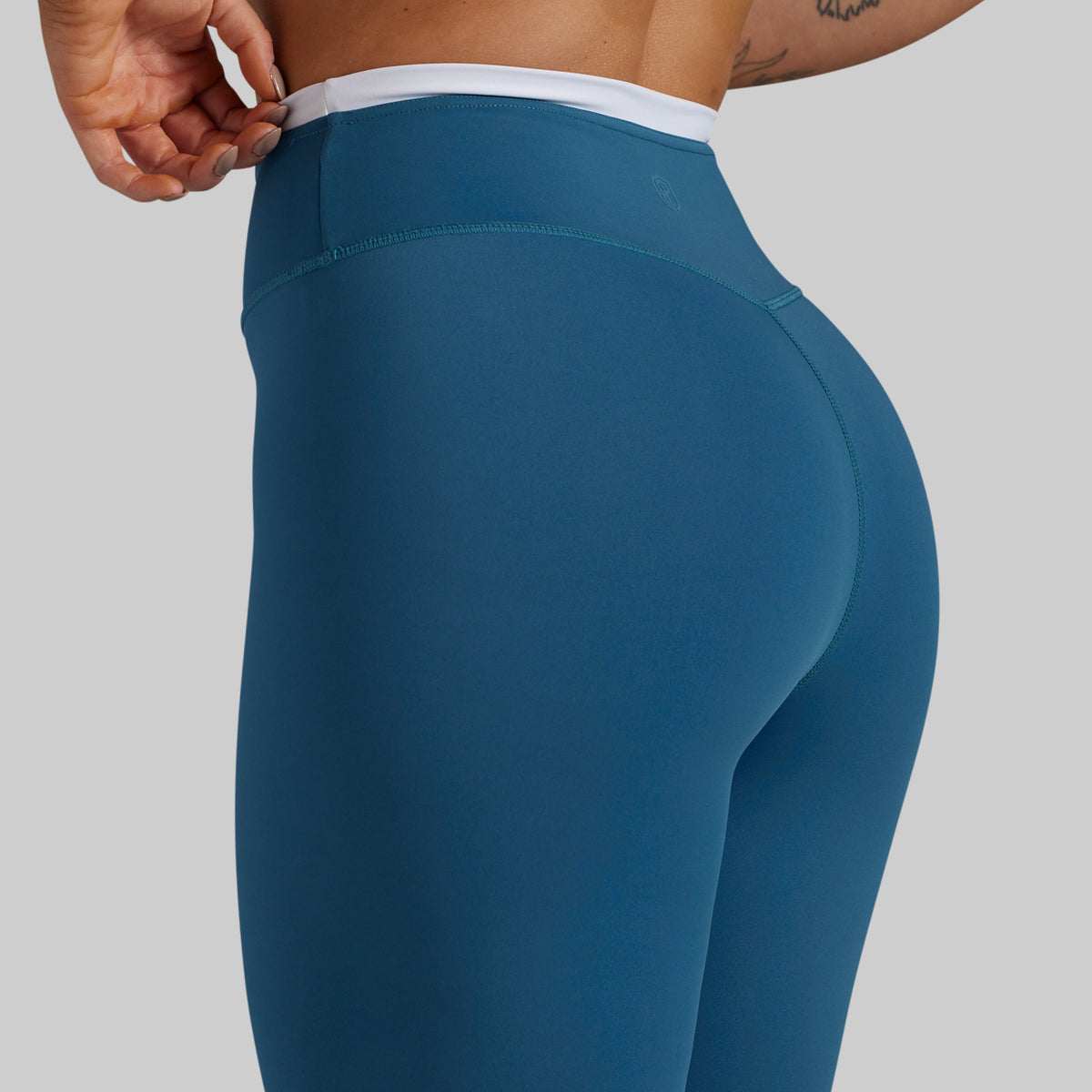 Close-up of a model wearing high-waisted teal blue leggings with a contrasting white waistband, shown from the back and side against a solid gray background.