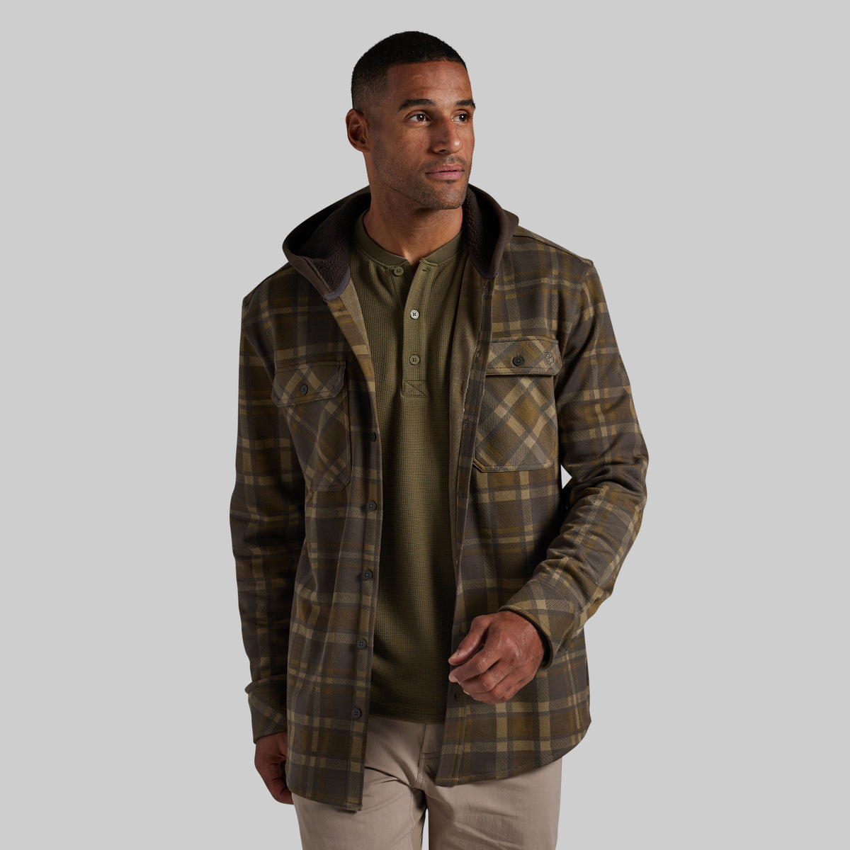 A man wearing a brown and olive green plaid hooded flannel over an olive green henley shirt and beige pants, photographed against a solid gray studio background.