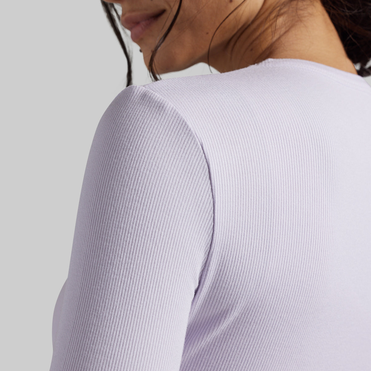 Close-up of a person wearing a light lavender ribbed long-sleeve top, showing the shoulder and upper back against a plain light gray background.