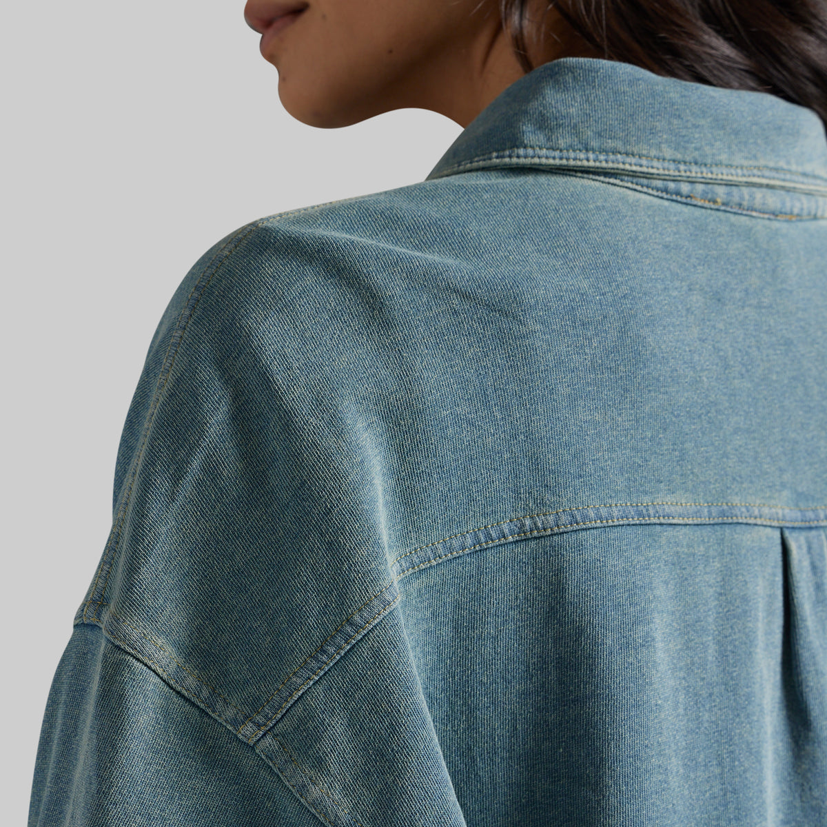 Close-up of a person wearing an oversized blue denim shirt, showing the back collar, shoulder, and denim texture against a light gray background.