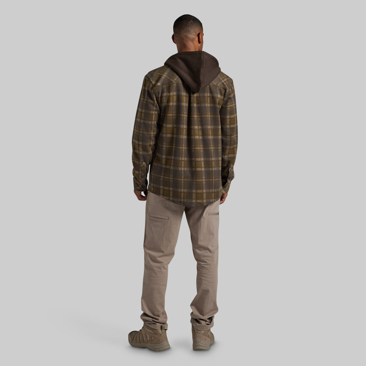 A man viewed from the back wearing a brown and olive plaid hooded flannel, tan pants, and brown boots against a solid gray studio background.
