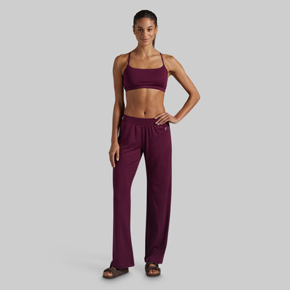 A woman modeling a matching two-piece loungewear set in deep burgundy, featuring a spaghetti-strap lounge bra and high-waisted wide-leg pants, set against a solid light gray background.