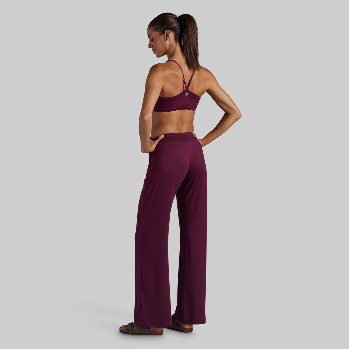 A woman stands in profile wearing a matching burgundy lounge set consisting of a lounge bra and high-waisted wide-leg pants.