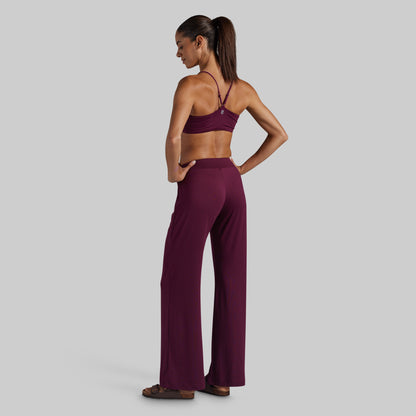 A woman stands in profile wearing a matching burgundy activewear set consisting of a sports bra and high-waisted wide-leg pants, photographed against a solid light gray background.