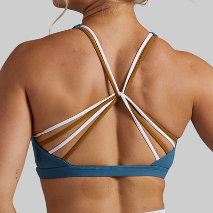 Rear view of a woman wearing a teal sports bra with multiple thin crossover straps in white and mustard brown on a light gray background.