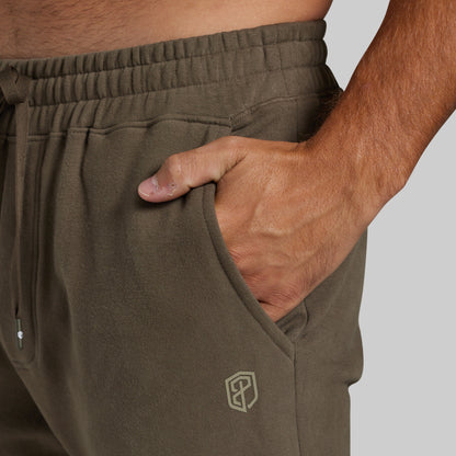 Close-up of a person wearing olive green sweatpants, with a hand tucked into the side pocket. The fabric has a soft texture, an elastic waistband with a drawstring, and a small geometric logo near the lower left leg.