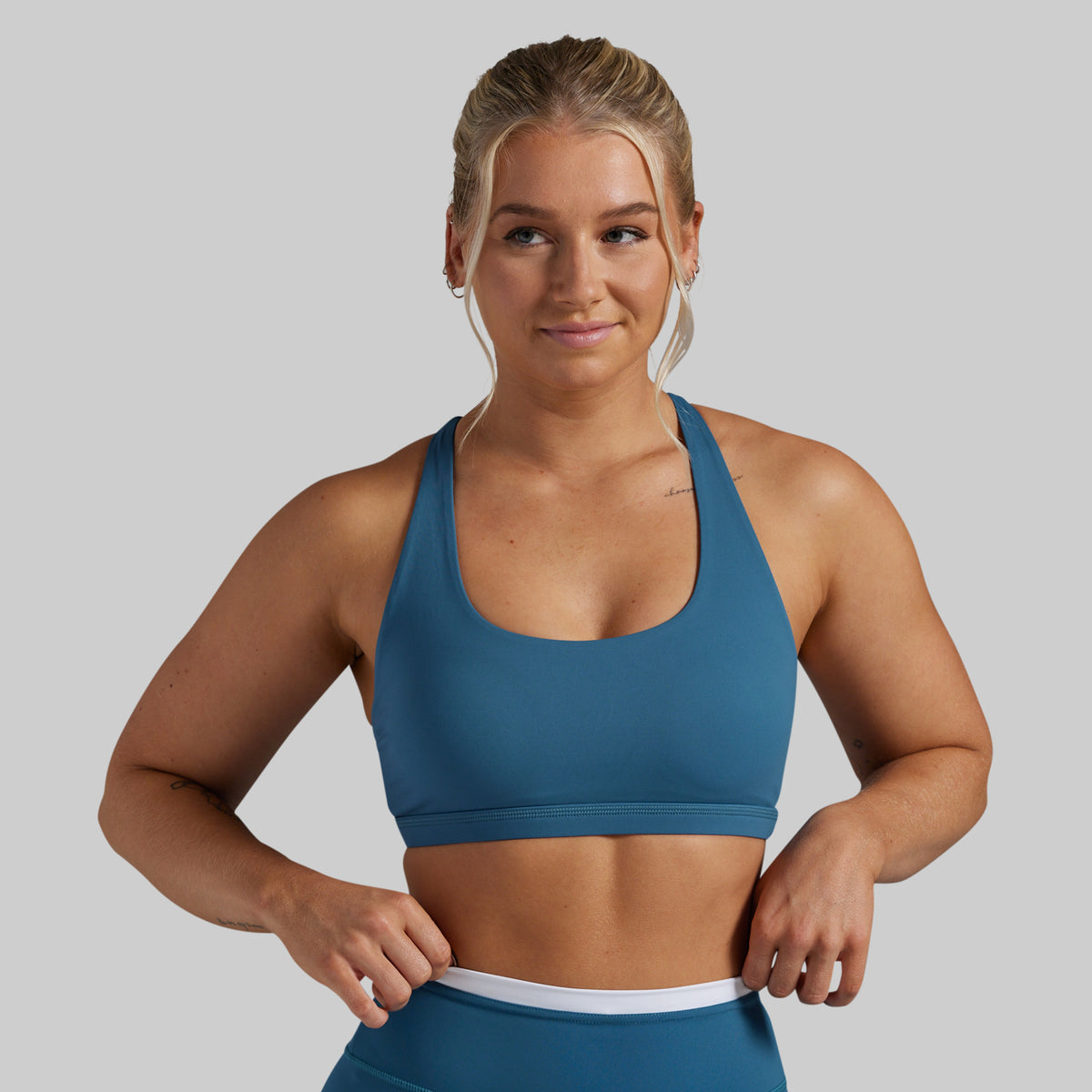 A woman modeling a slate blue athletic sports bra with a wide scoop neckline and a white waistband, paired with matching blue athletic bottoms, against a solid gray background.