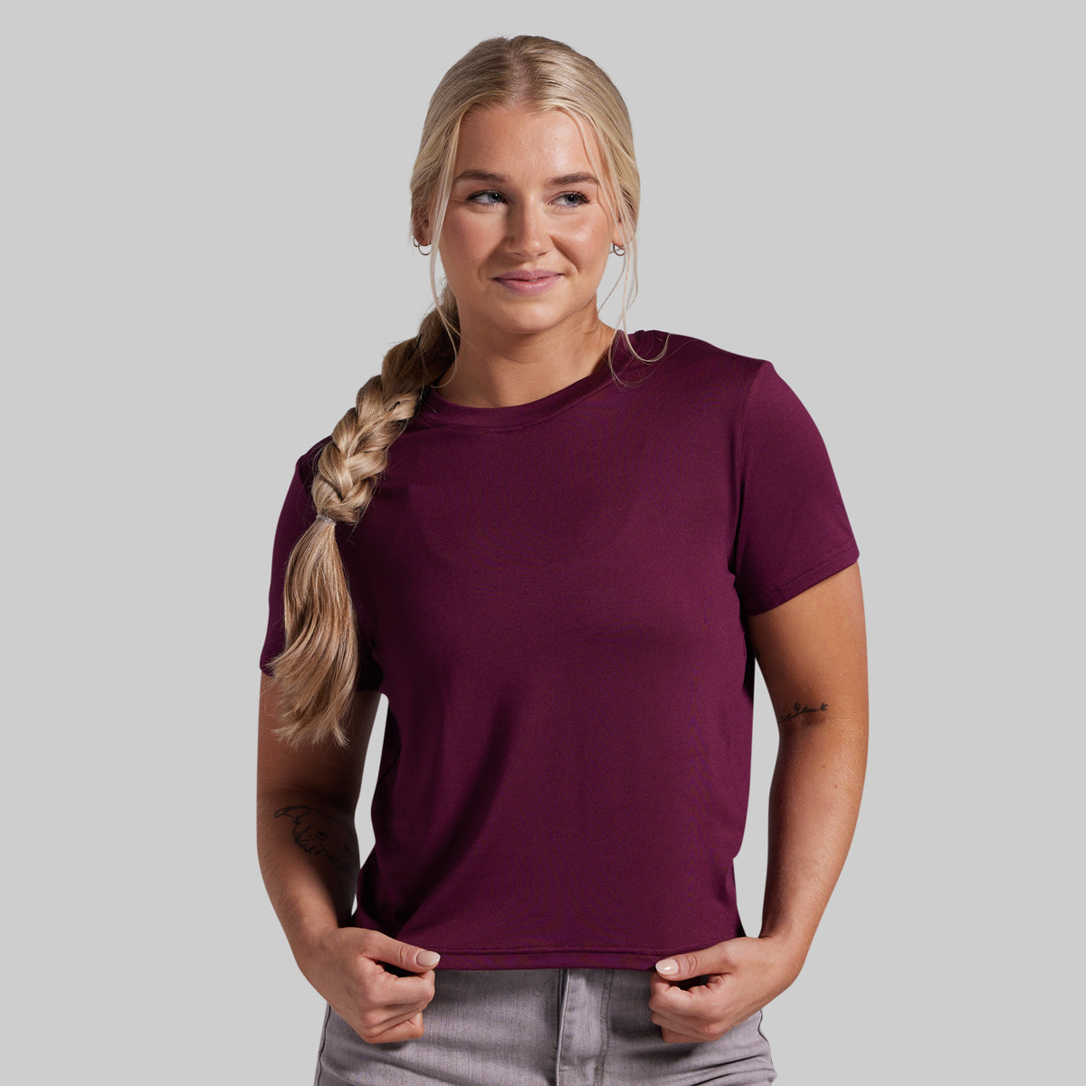 A woman with blonde hair in a side braid wearing a short-sleeve crewneck t-shirt in deep burgundy, paired with gray pants, against a solid gray background.