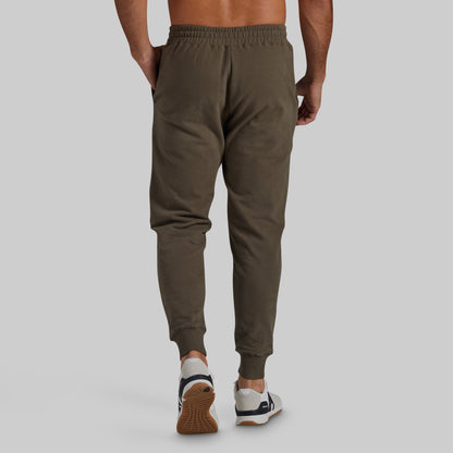 A man viewed from behind wearing olive green jogger sweatpants and white sneakers with brown soles, set against a solid light gray background.