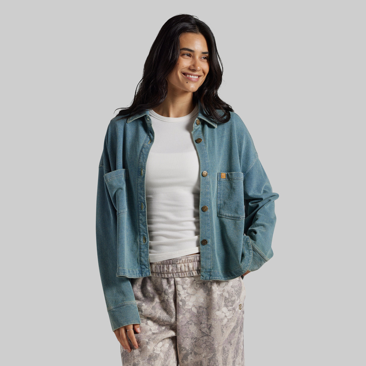 A woman modeling a light blue denim button-up shirt jacket over a white t-shirt and patterned pants, photographed against a solid gray studio background.