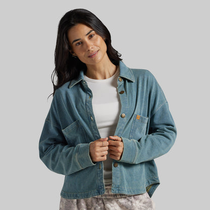 A woman wearing a light blue denim button-up shirt jacket over a white ribbed top, photographed against a solid light gray studio background.
