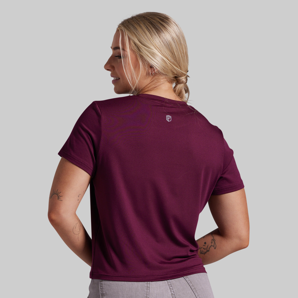 A woman shown from the back wearing a short-sleeve athletic t-shirt in deep burgundy, paired with light gray leggings, against a solid light gray background.