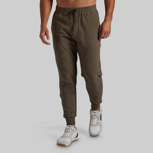 A man wearing olive green jogger sweatpants with a drawstring waist and elastic cuffs, paired with white sneakers, shown from the waist down against a light gray studio background.