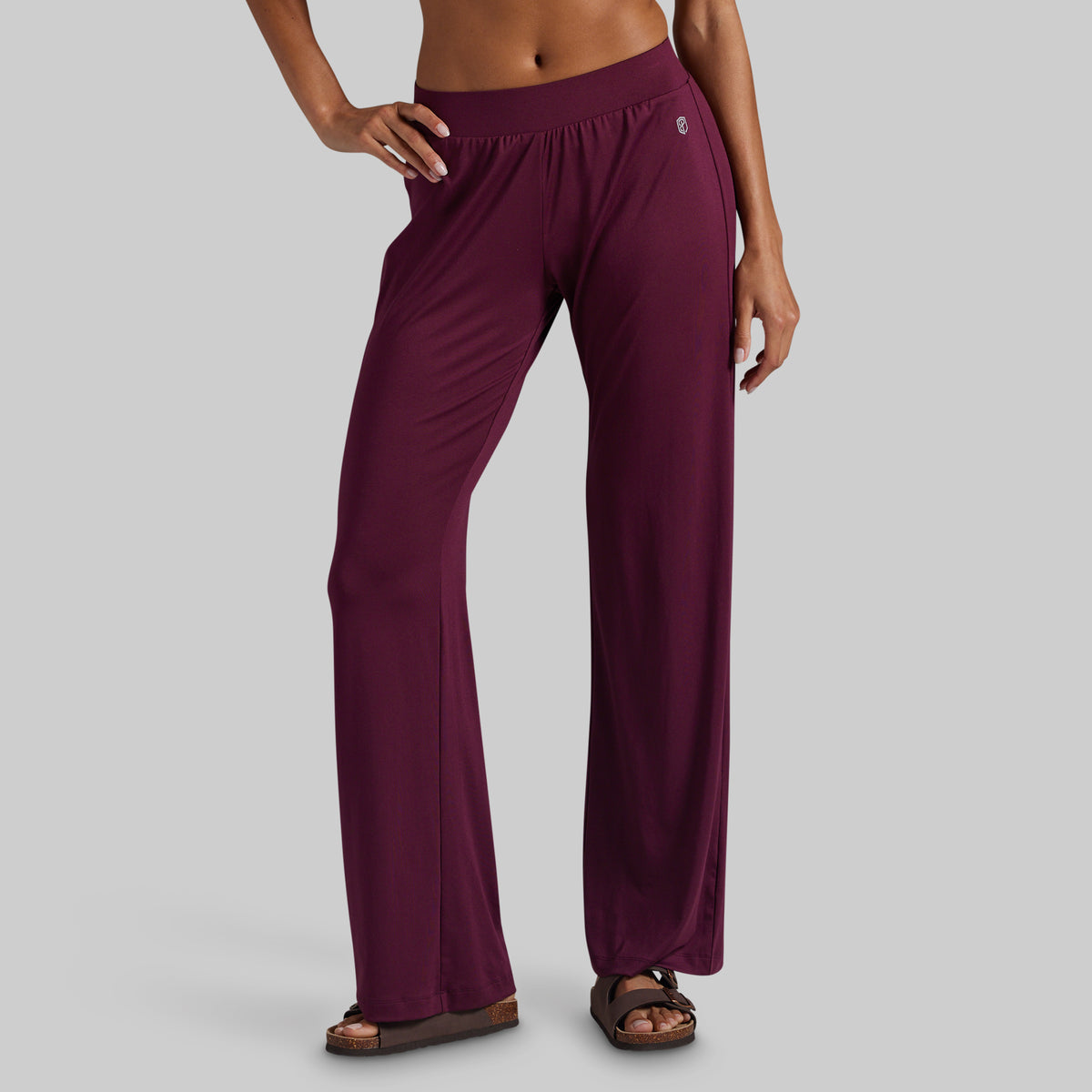 A model wearing a pair of wide-leg, high-waisted lounge pants in deep burgundy, paired with brown sandals, shown from the waist down against a solid gray background.