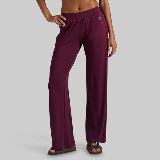 A model wearing a pair of wide-leg, high-waisted lounge pants in deep burgundy, paired with brown sandals, shown from the waist down against a solid gray background.