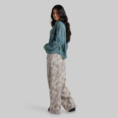 A woman modeling a teal cropped denim jacket and wide-leg beige and gray patterned pants against a solid light gray background.