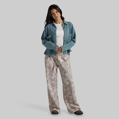 A woman modeling a casual outfit consisting of a teal cropped denim jacket over a white top and wide-leg beige and gray patterned pants, set against a solid gray studio background.