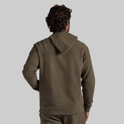A man viewed from behind wearing an olive brown hooded sweatshirt and matching sweatpants against a solid gray background.