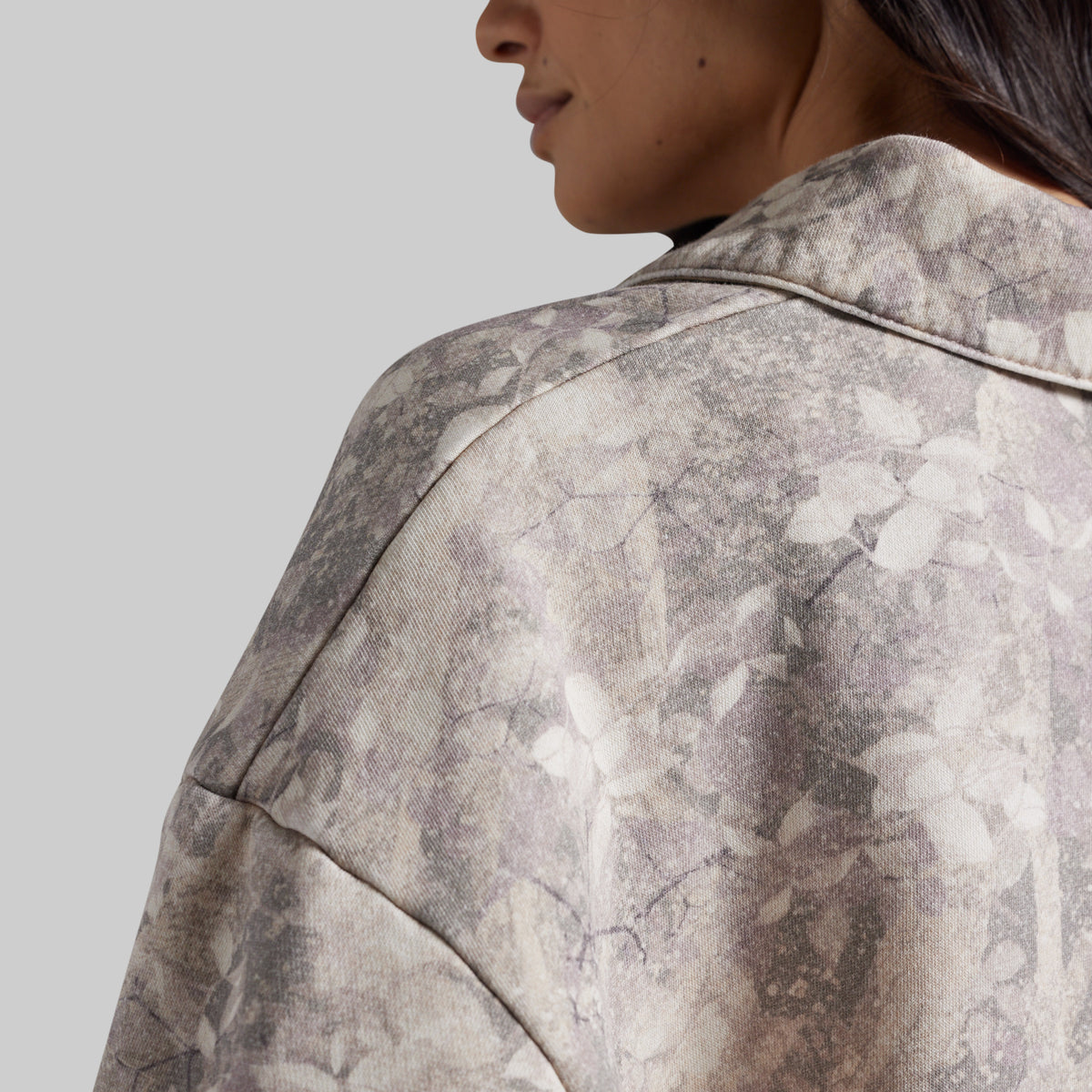 Close-up of a person wearing an abstract-patterned sweatshirt in muted gray, beige, and off-white tones, shown from the side against a plain light gray background.