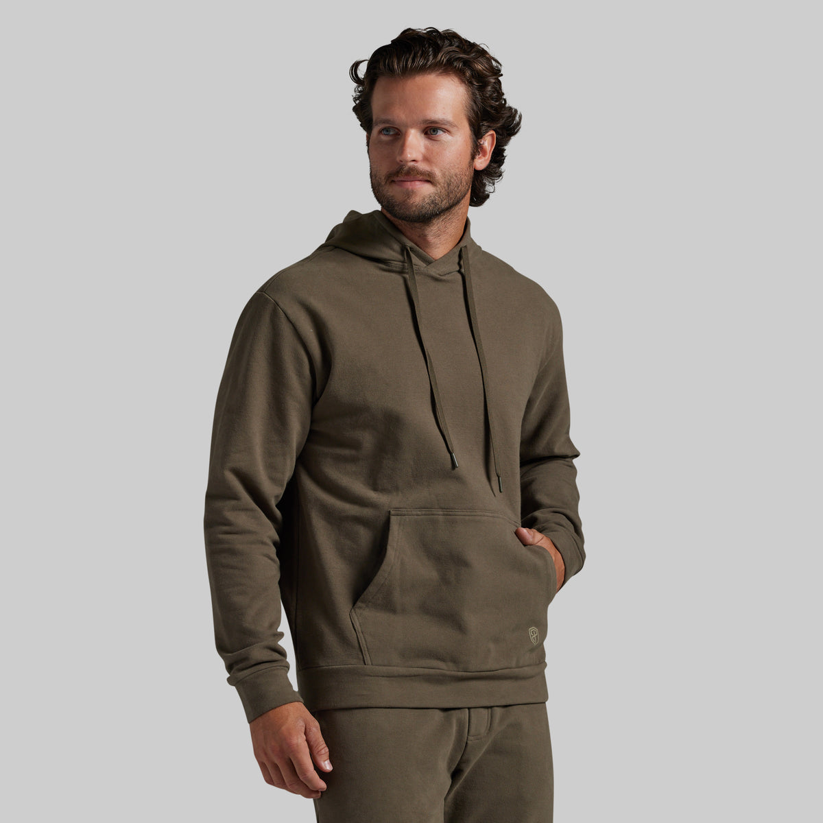 A man wearing an olive green pullover hoodie with matching sweatpants, standing against a solid light gray background.