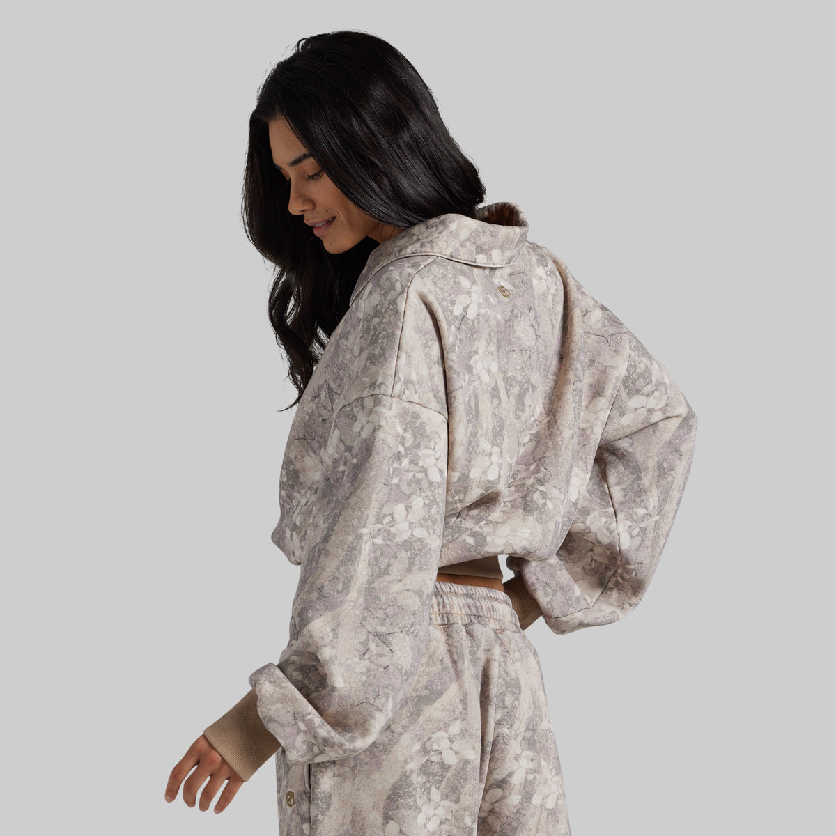 A woman wearing a matching cropped sweatshirt and sweatpants set in a beige and gray abstract floral pattern, shown from a side-back angle against a solid gray studio background.