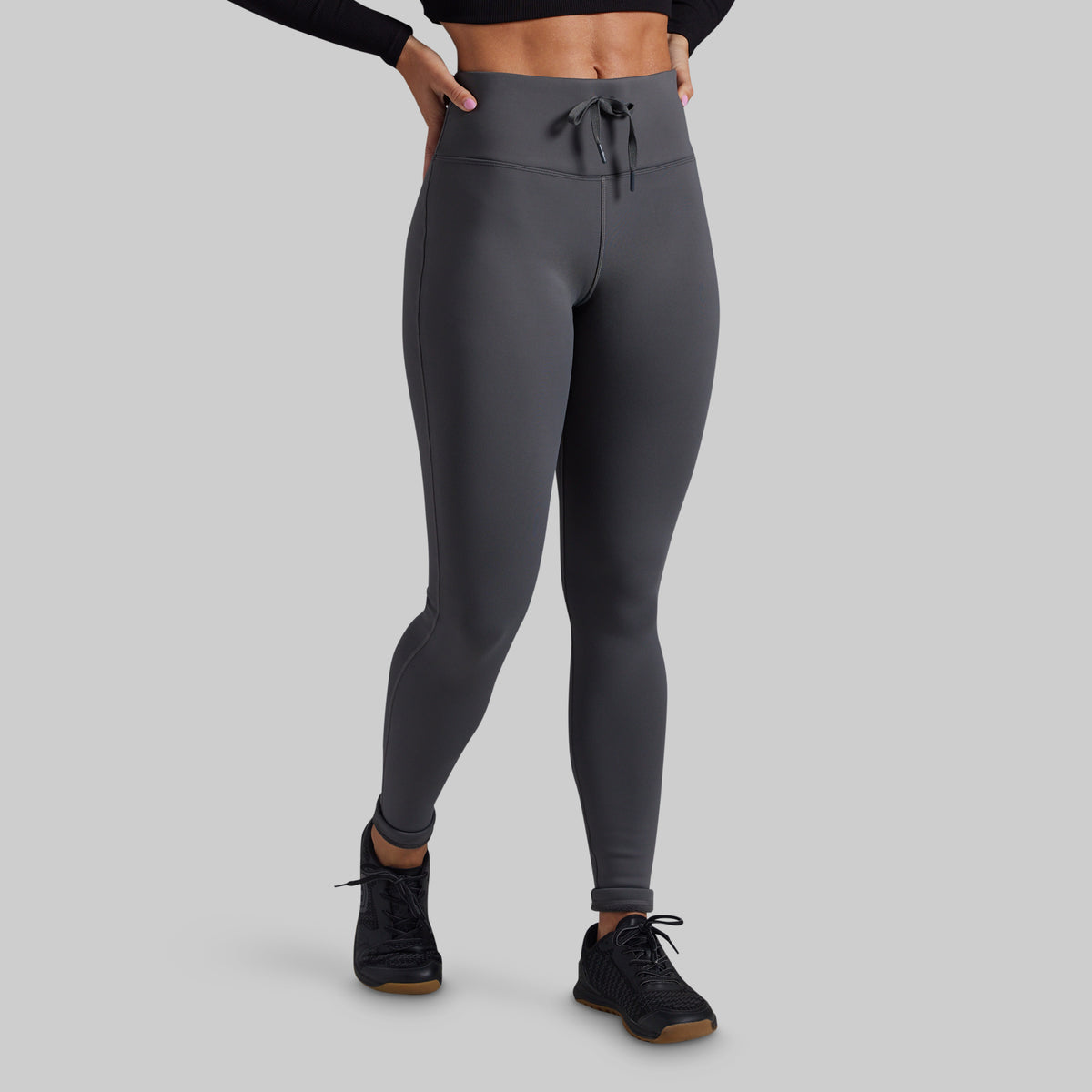 A model wearing dark gray high-waisted athletic leggings with a drawstring waistband, paired with black athletic shoes, shown from the waist down on a solid gray background.