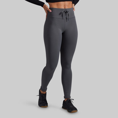 A model wearing dark gray high-waisted athletic leggings with a drawstring waistband, paired with black athletic shoes, shown from the waist down on a solid gray background.