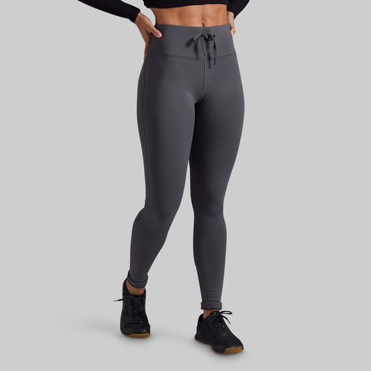 A model wearing dark gray high-waisted athletic leggings with a drawstring waistband, paired with black athletic shoes, shown from the waist down on a solid gray background.