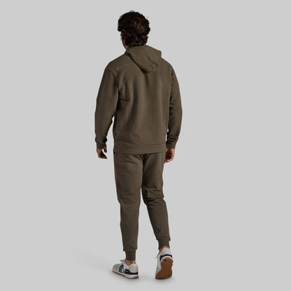 A man viewed from behind wearing an olive green hooded sweatshirt and matching jogger pants, paired with white and brown sneakers, against a solid light gray background.