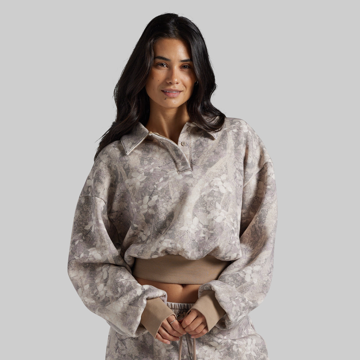 A woman modeling a cropped, long-sleeve collared sweatshirt with a mottled beige and gray camouflage pattern, paired with matching beige drawstring pants, against a solid gray studio background.