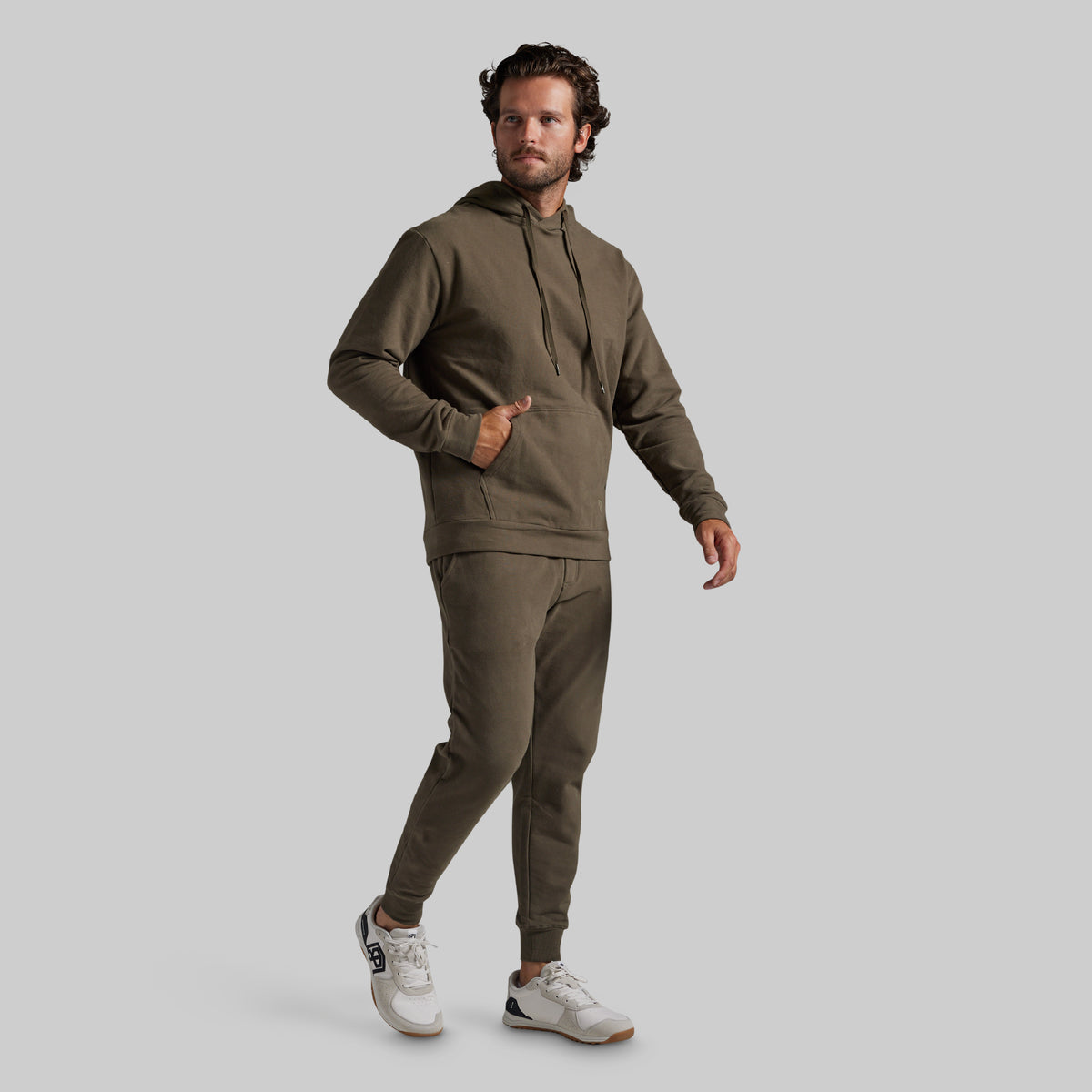 A man modeling a matching olive green hoodie and jogger sweatpants set, paired with white sneakers, against a solid light gray background.