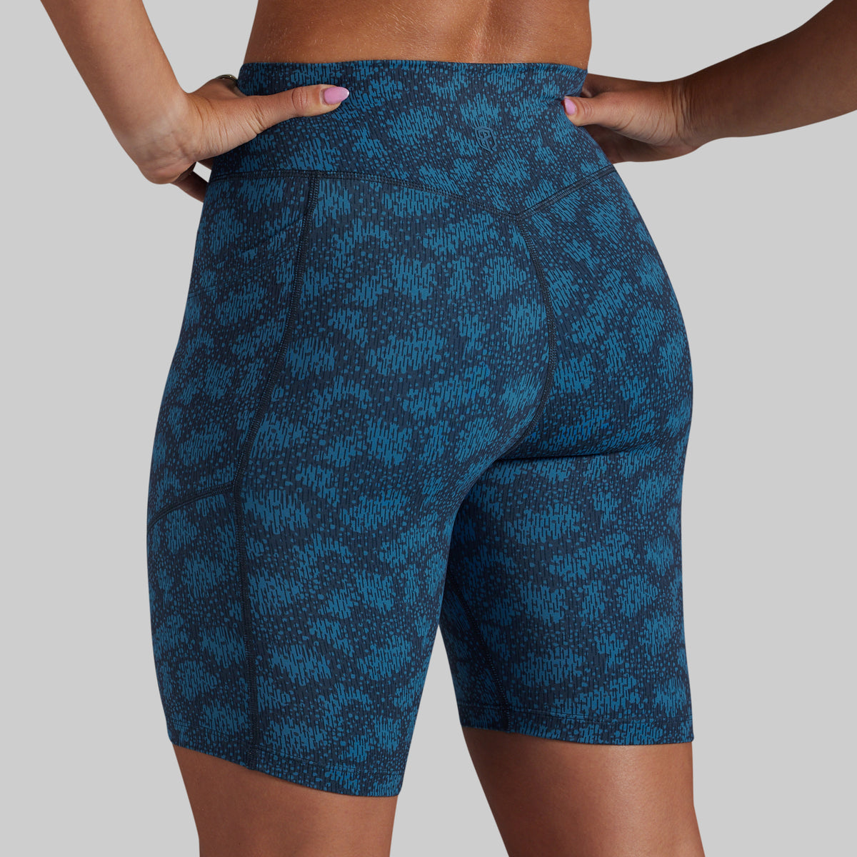 A close-up of a model wearing high-waisted blue and black patterned biker shorts, shown from the back against a solid gray background.