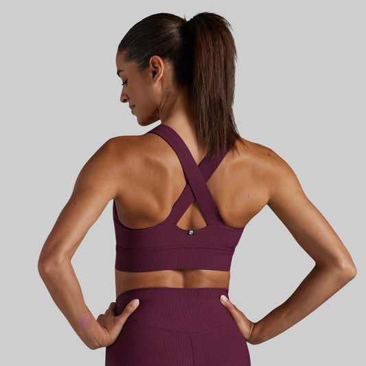 A woman shown from the back wearing a ribbed athletic sports bra and matching high-waisted leggings in a deep plum purple color, set against a solid light gray background.
