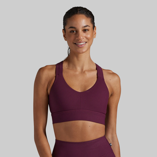 A woman modeling a ribbed athletic sports bra in a deep plum purple color, paired with matching high-waisted leggings, against a solid light gray background.