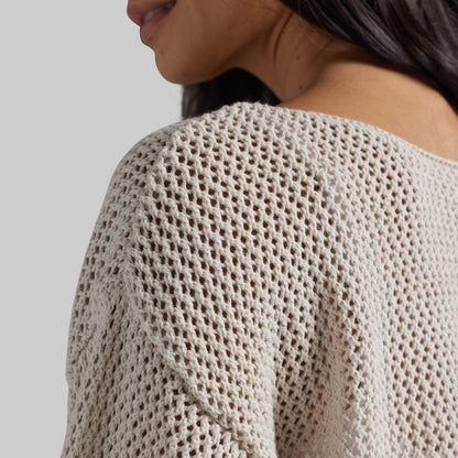 Close-up of a person wearing a cream-colored open-knit sweater, showing the textured knit pattern and shoulder detail against a light gray background.