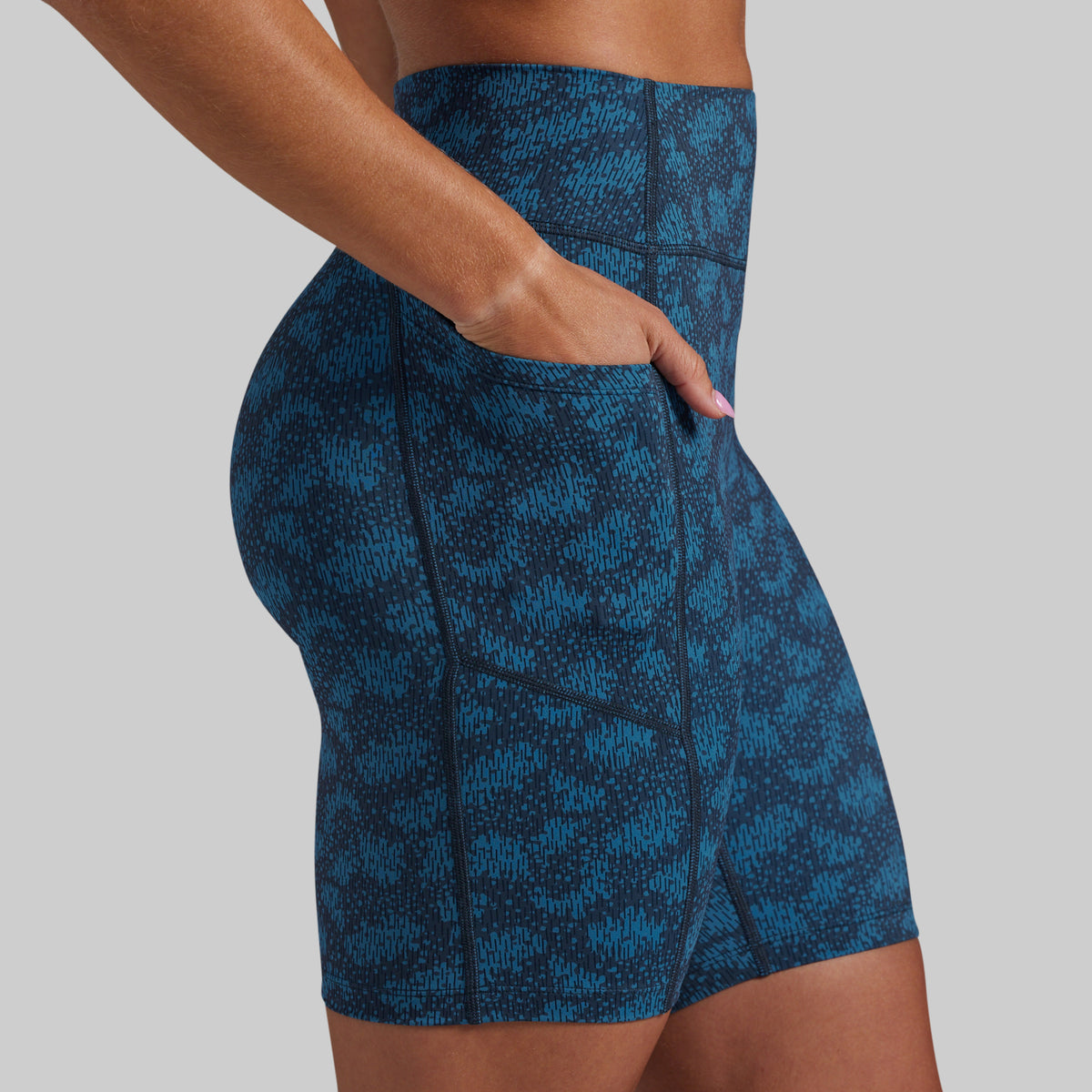 Close-up side view of a model wearing high-waisted athletic shorts with a blue and black abstract pattern and a side pocket, against a light gray background.