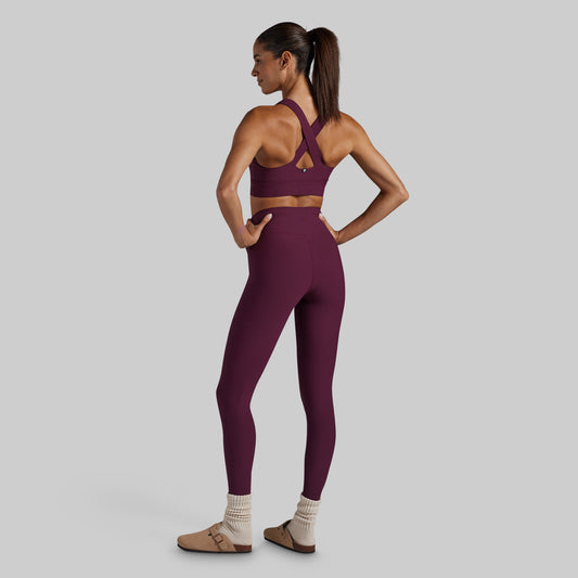 A woman modeling a matching burgundy activewear set, featuring a sports bra and high-waisted leggings, shown from a rear three-quarter view against a solid gray background.