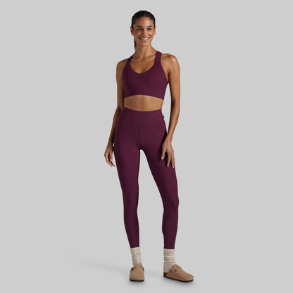 A woman modeling a matching athletic set consisting of a scoop-neck sports bra and high-waisted leggings in a deep plum purple color, styled with beige socks and tan shoes against a solid gray background.