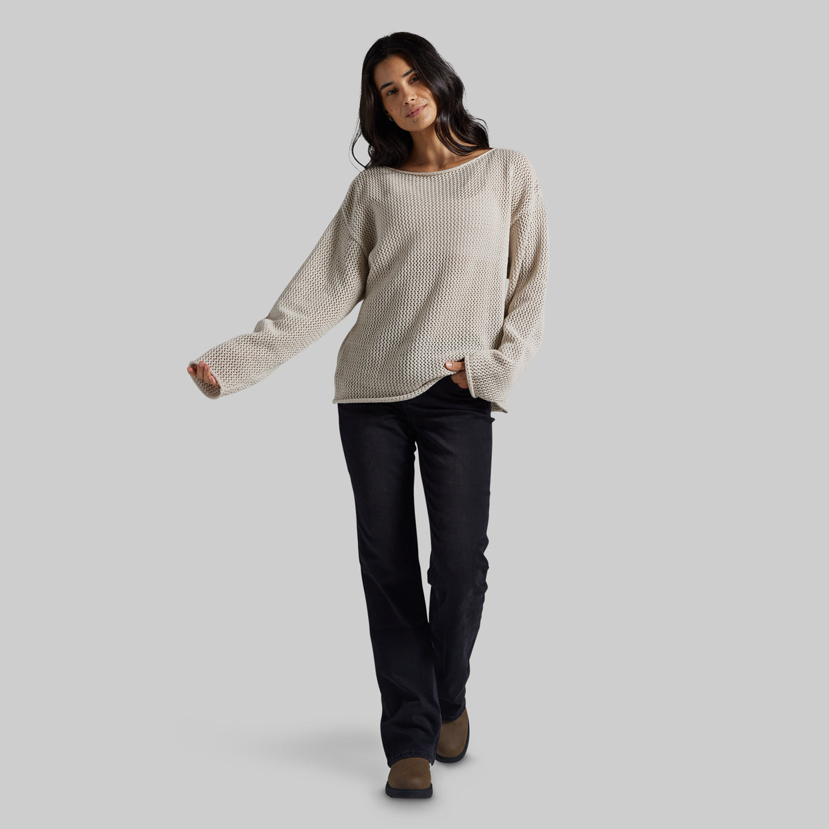 A woman modeling a cream-colored, long-sleeve waffle-knit sweater with dark pants, standing against a solid light gray background.