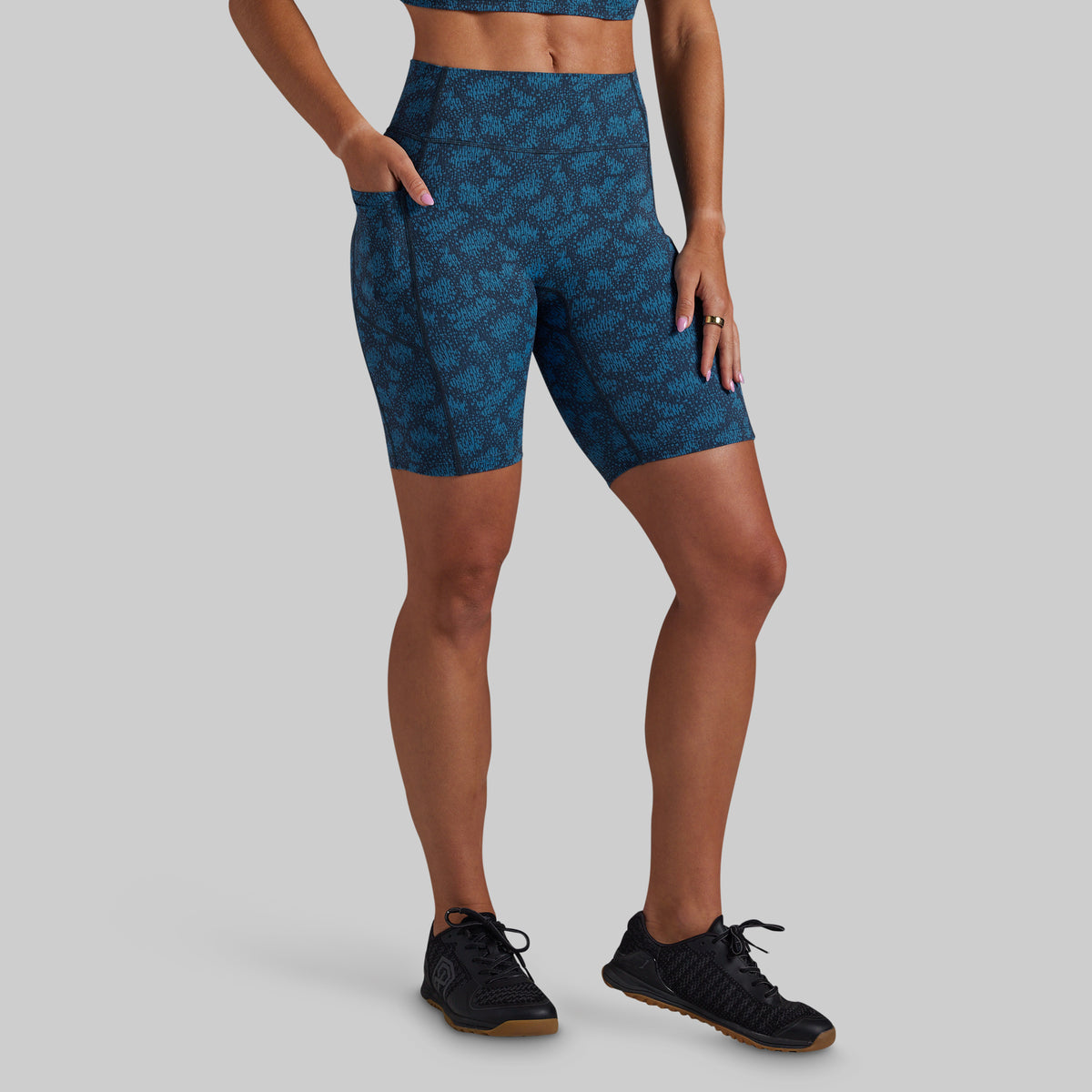 A model wearing high-waisted blue athletic biker shorts with a tonal abstract pattern, paired with black sneakers, shown against a solid gray background.