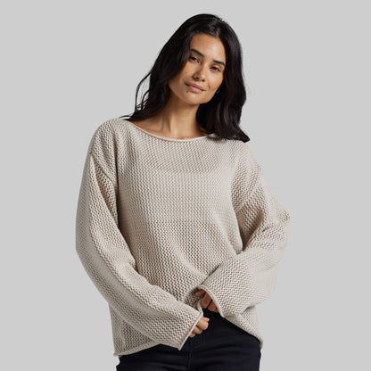 A woman wearing a textured, oversized knit sweater in a light beige color, paired with black pants, against a solid light gray background.