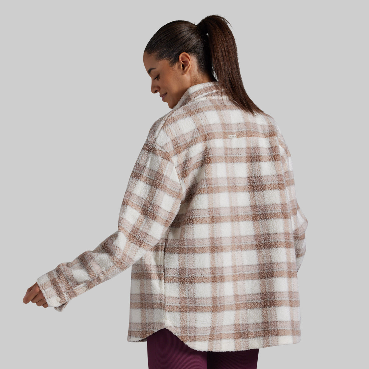 A woman wearing an oversized, long-sleeve plaid fleece button-up shirt in beige and white, shown from a three-quarters back view against a solid gray background.